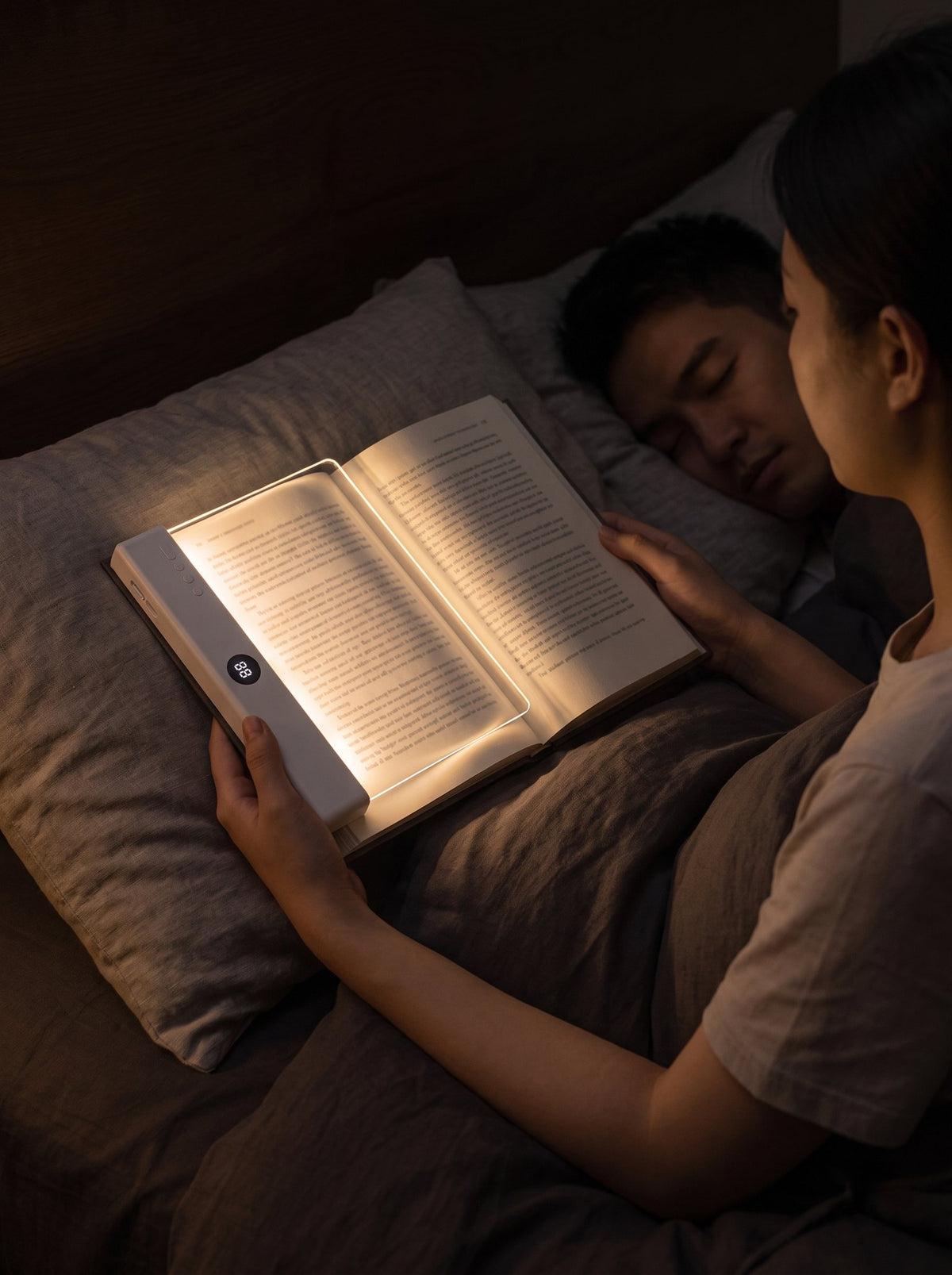 A woman reads in bed by the glow of the Serene Light, which lies on her open book, while her partner sleeps peacefully beside her.